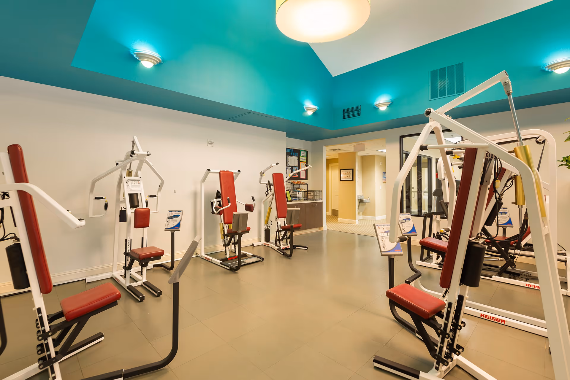 A fitness room with several exercise machines featuring red seats and white frames. The room has a teal ceiling with wall-mounted lights and a large overhead light. There is a mirror on the right wall and an open doorway leading to a hallway.