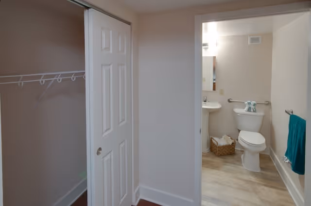 View of a small bathroom with a white toilet, pedestal sink, and a mirror above the sink. There is a grab bar behind the toilet and a teal towel hanging on a towel rack to the right. A basket with towels is placed on the floor next to the toilet. To the left of the bathroom is an open closet with a white sliding door and a hanging rod with hooks.