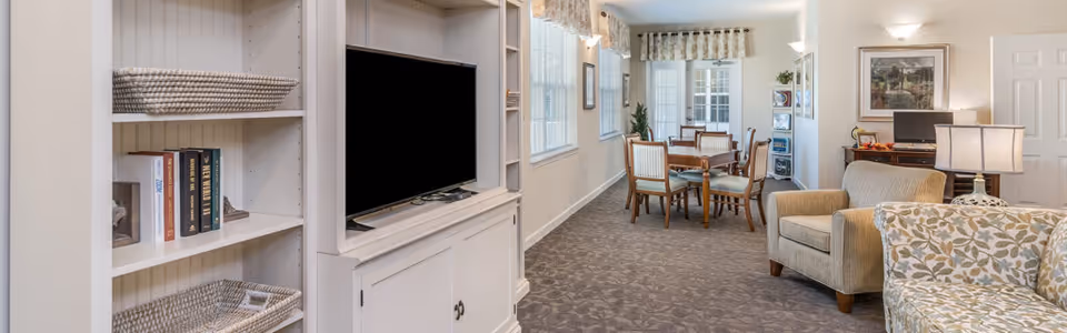 A bright and spacious living area featuring a white entertainment center with a flat-screen TV, built-in shelves with books and baskets, a beige armchair, a floral patterned sofa, a wooden dining table with chairs, and large windows letting in natural light.