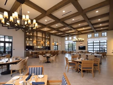 Spacious dining room with wooden tables and chairs, large windows letting in natural light, decorative chandeliers hanging from a ceiling with exposed wooden beams, and a bar area with pendant lighting in the background.