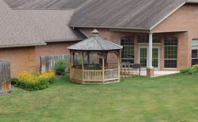 A grassy backyard area with a wooden gazebo in the center, surrounded by a wooden fence and yellow flowers along the fence line. In the background, there is a brick building with large windows and a covered patio area.