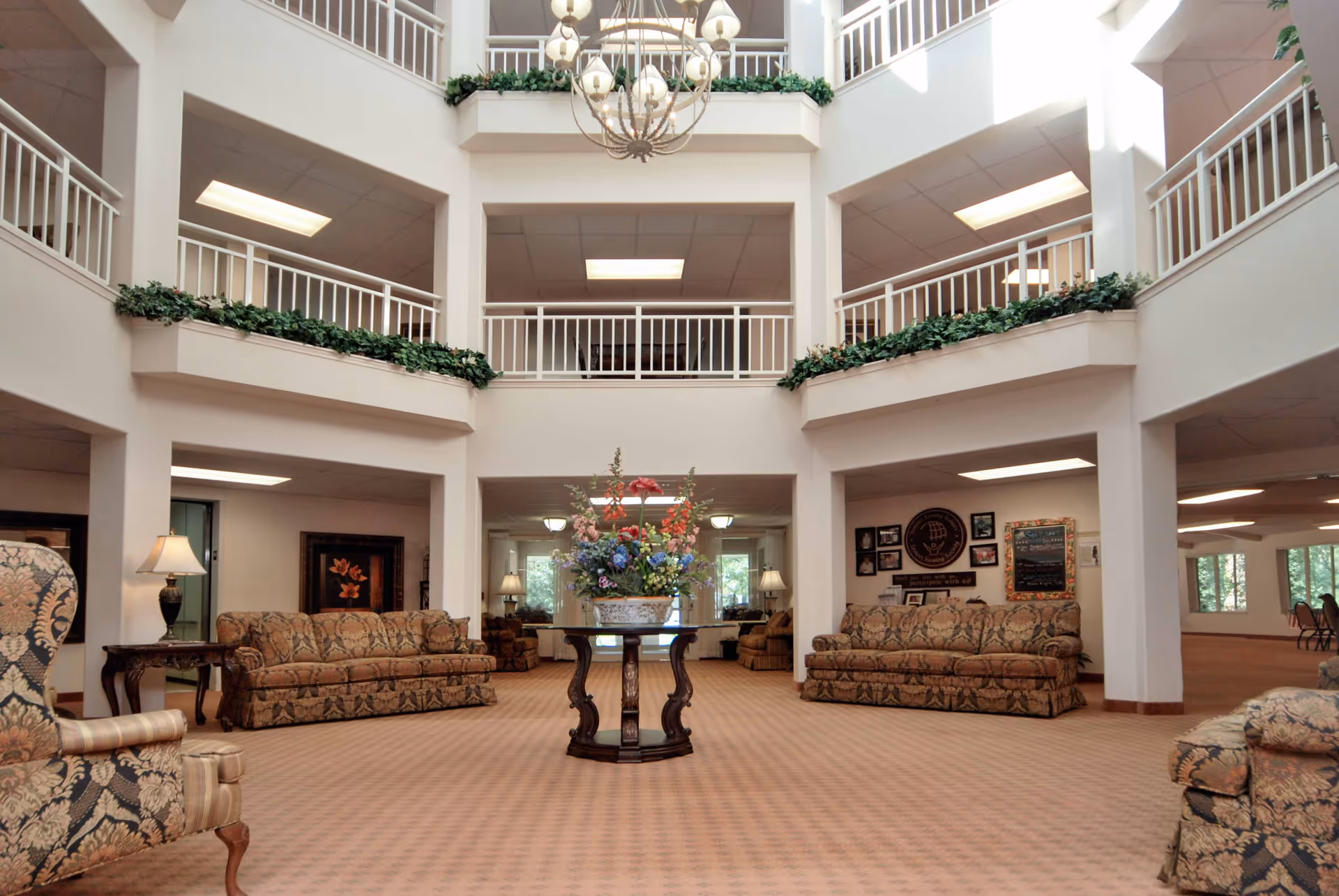 Spacious and well-lit interior common area of a senior living facility with patterned sofas, armchairs, a central round table with a floral arrangement, and a high ceiling with balconies and hanging chandeliers.