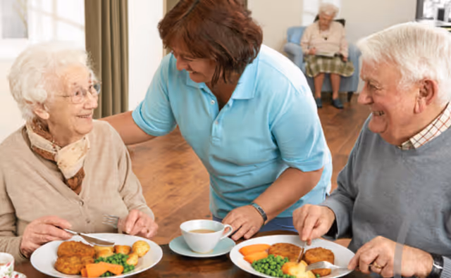 Caregiver leaning over and serving meals to two smiling elderly residents seated at a dining table with plates of vegetables and a cup of tea.
