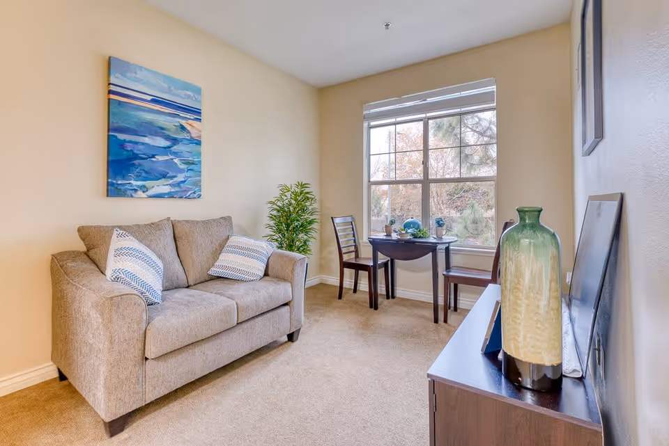 A cozy living room area with a beige loveseat adorned with two patterned pillows, a small dark wooden table with two chairs placed near a large window showing trees outside, a green and beige decorative vase on a dark wooden cabinet, and a colorful abstract painting on the wall.