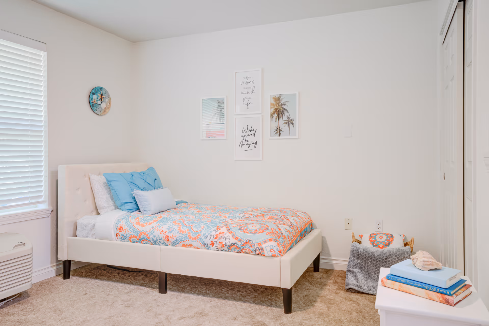 A bright and cozy bedroom with a single bed featuring a white upholstered headboard and colorful patterned bedding in shades of orange and blue. The bed has multiple pillows in blue and white. On the wall above the bed are four framed pictures, including palm trees and inspirational quotes. To the right of the bed is a small white nightstand with a stack of books and a decorative shell on top. A basket with a blanket and pillow sits on the floor next to the nightstand. The room has beige carpet, white walls, a window with white blinds, and a white closet door.