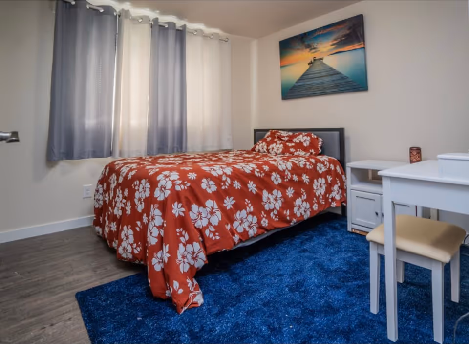 A bedroom with a bed covered in a red floral bedspread, a blue rug underneath, a white desk with a beige chair, a small white nightstand, and a wall painting of a pier extending into a calm body of water at sunset. The room has light-colored walls and two windows with gray curtains.