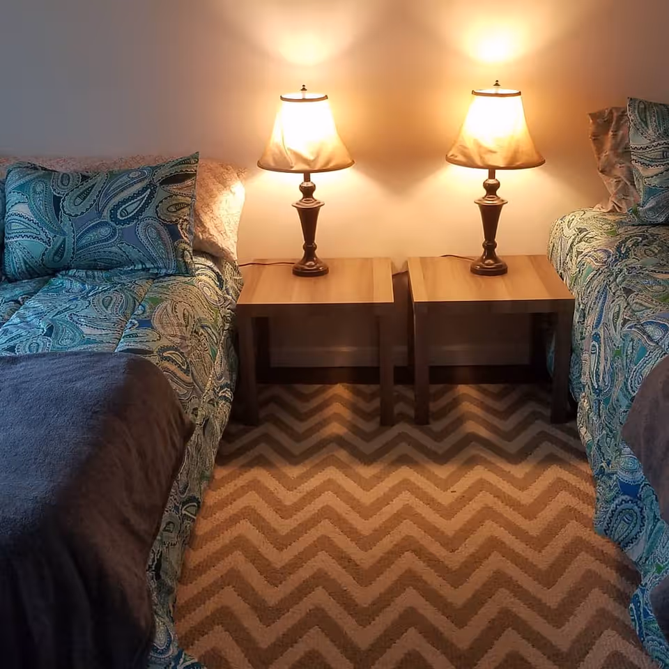 Two twin beds with blue and green patterned bedding separated by two wooden nightstands, each with a lit table lamp, on a chevron-patterned carpeted floor.