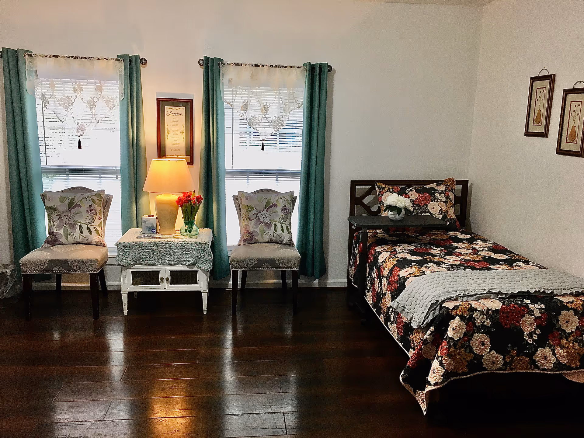 A cozy bedroom with a single bed covered in a floral patterned bedspread and a gray blanket. Next to the bed is a small table with a vase of white flowers. Two chairs with floral cushions flank a small white table with a lamp and a vase of red tulips. The room has two windows with teal curtains and lace valances, and three framed pictures hang on the wall above the bed.