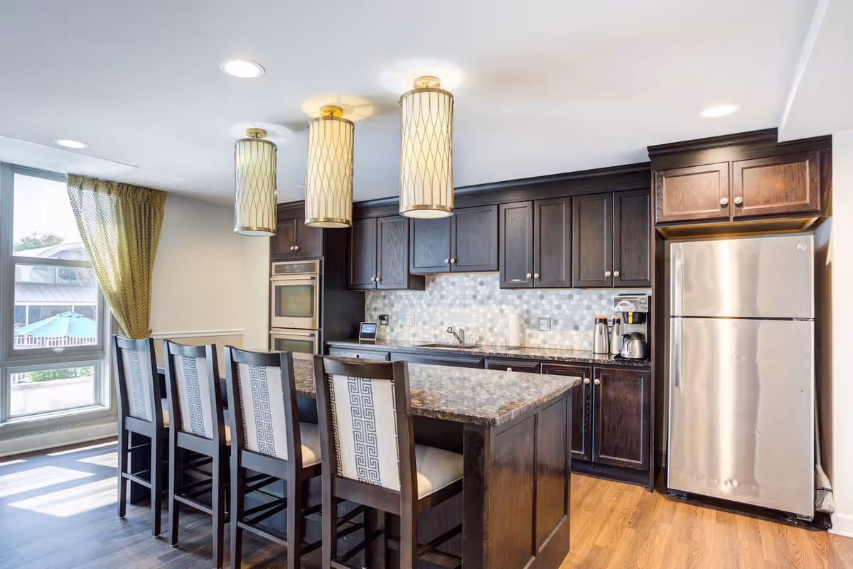 Modern kitchen with dark wood cabinets, a large island with granite countertop, four high-back chairs, stainless steel refrigerator, double oven, and three pendant lights hanging from the ceiling. A large window with green curtains allows natural light to fill the room.