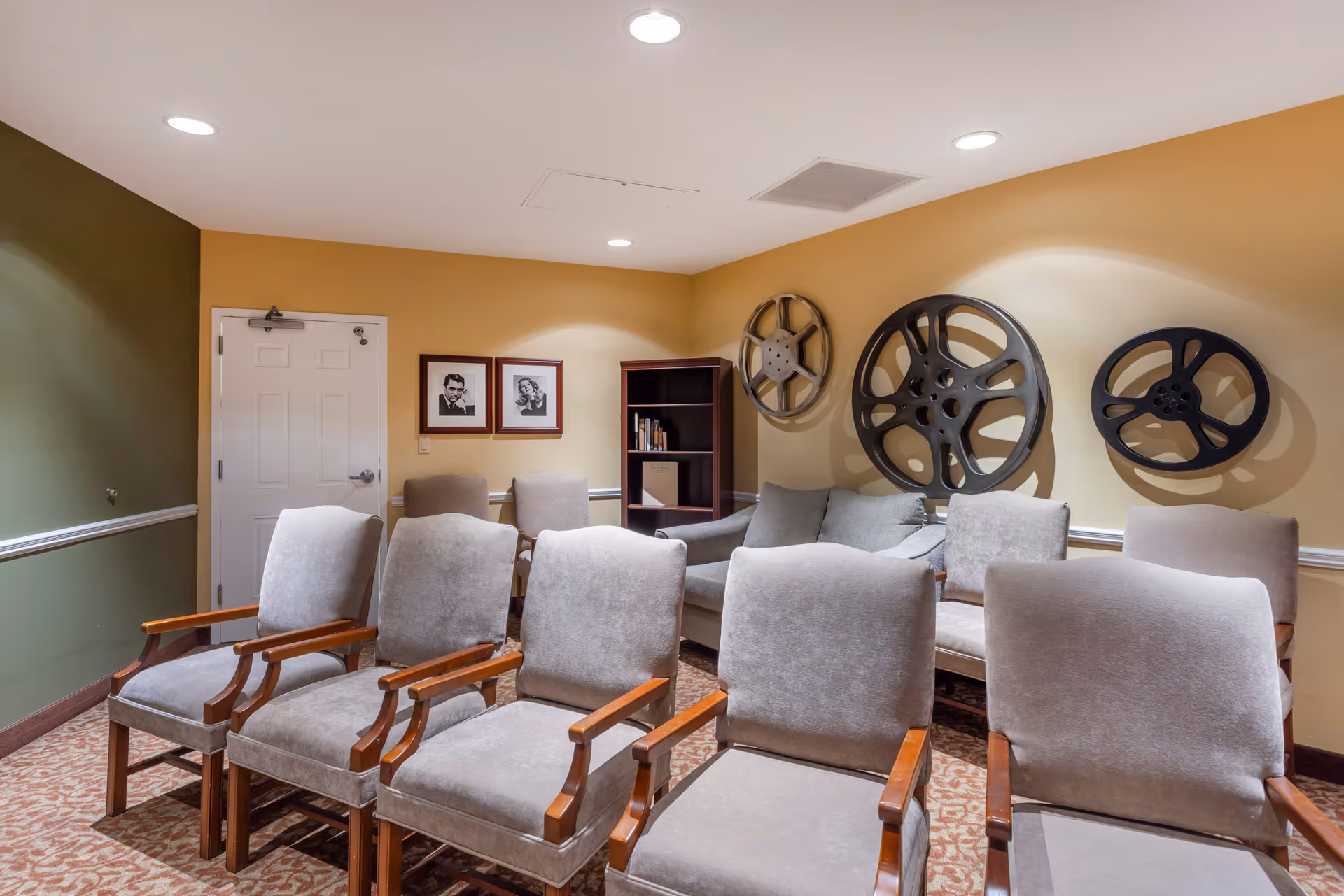 A small theater room with two rows of beige cushioned chairs with wooden armrests facing forward. The walls are painted yellow and green, decorated with three large vintage film reels and two framed black and white portraits. There is a gray couch and a small bookshelf in the back corner. The room has recessed ceiling lights and patterned carpet flooring.
