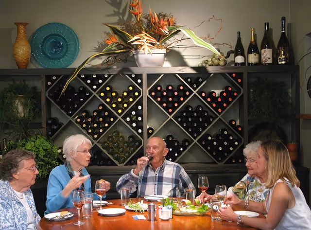 Five elderly people sitting around a dining table enjoying drinks and conversation. Behind them is a wine rack filled with bottles and a decorative arrangement of flowers and plants on top.