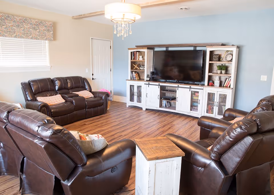 A cozy living room with four dark brown leather recliners arranged around a small wooden side table. Against a light blue wall is a large white entertainment center with shelves holding books, plants, and decorative items, and a flat-screen TV in the center. The room has wood flooring, a window with a patterned valance, and a chandelier hanging from the ceiling.