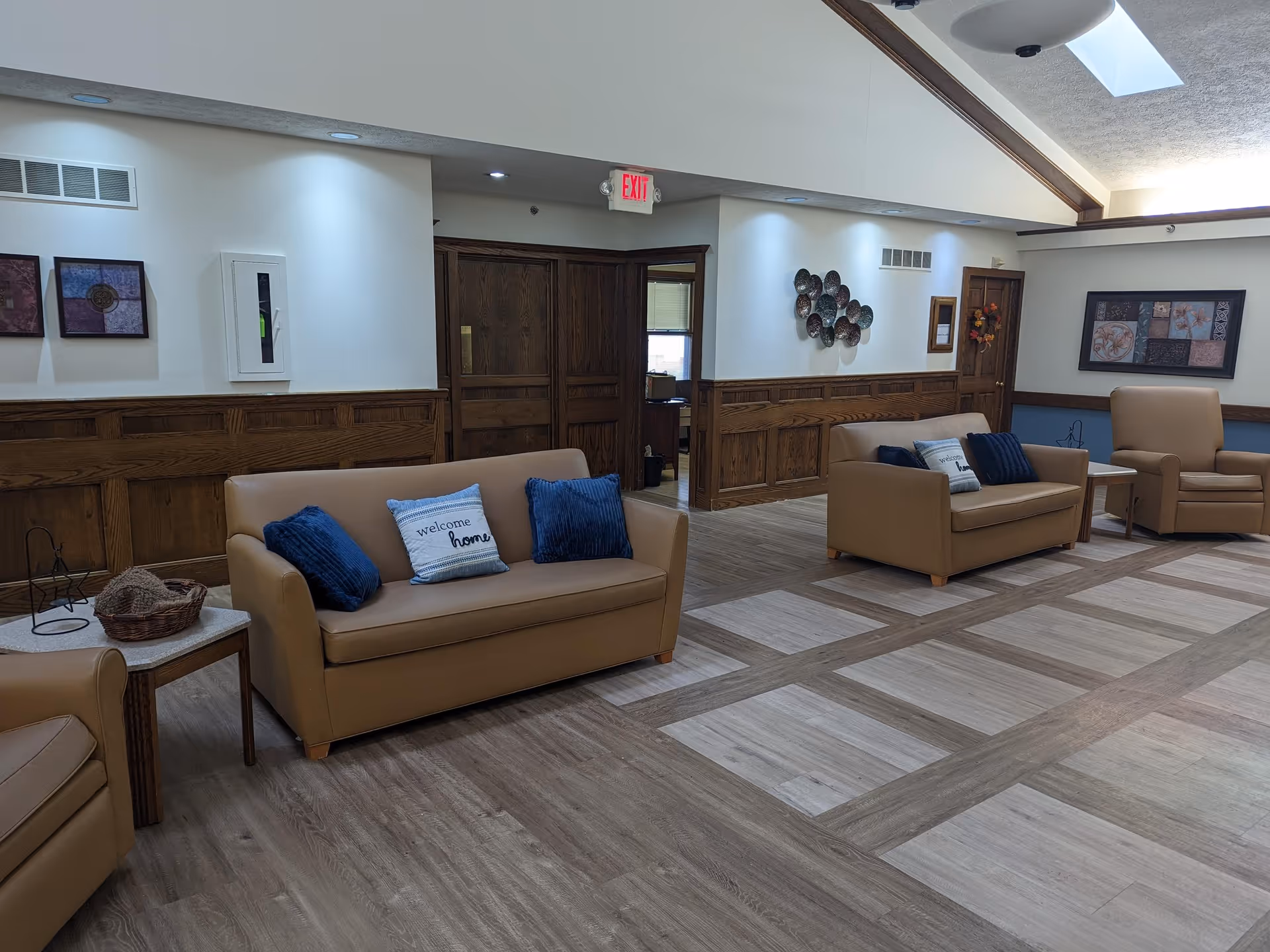 A spacious senior living facility common area with beige leather sofas and armchairs, each adorned with blue and white pillows. The room features wood paneling on the walls, decorative wall art, and a patterned wood floor. There is an exit sign above a set of wooden double doors, and the ceiling has skylights allowing natural light to enter.