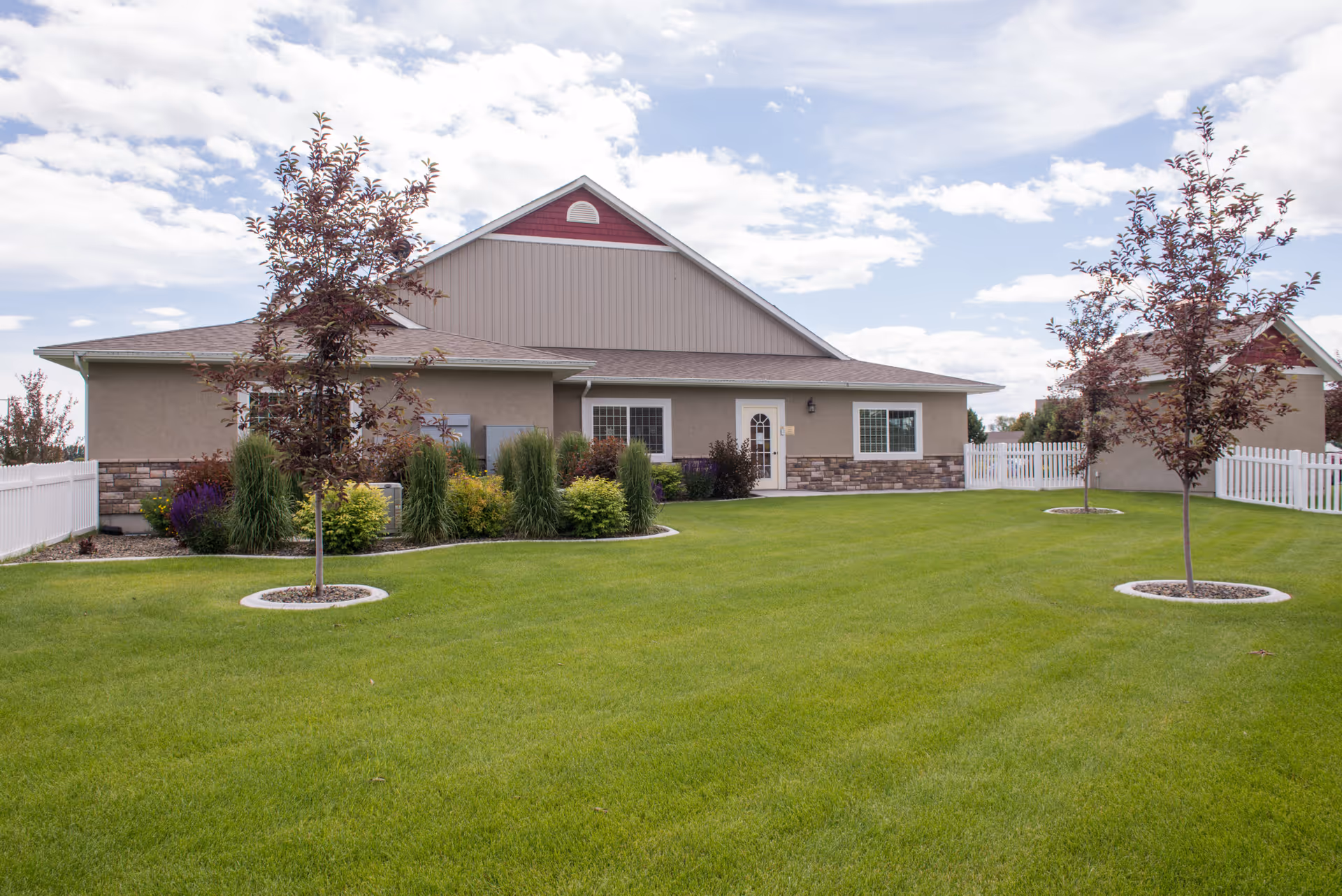 Exterior view of The Gables Assisted Living of Blackfoot building with a well-maintained green lawn, small trees planted in circular stone beds, and a white picket fence surrounding the property under a partly cloudy sky.