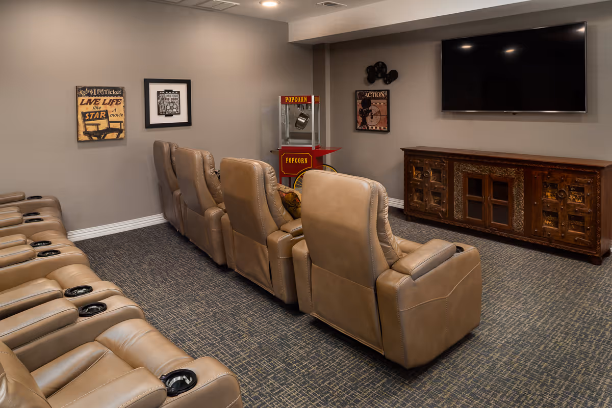 A cozy senior living facility media room with two rows of beige leather recliner chairs facing a large flat-screen TV mounted on a gray wall. There is a vintage-style popcorn machine in the corner and decorative movie-themed wall art. The room has carpeted flooring and recessed ceiling lights.