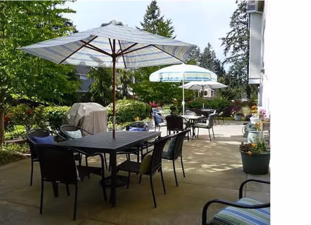 Outdoor patio area with multiple tables and chairs, each table shaded by large umbrellas. The patio is surrounded by greenery including trees and bushes, with a barbecue grill visible in the background. The setting appears to be a relaxing outdoor space at Bridgeport Place Assisted Living.