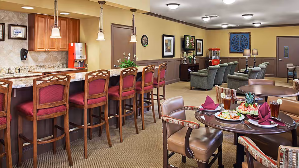 Interior view of a senior living facility common area featuring a long counter with six wooden bar stools with red cushions, a kitchen area with cabinets and a beverage dispenser, and a dining area with round tables set with plates of food, drinks, and folded napkins. In the background, there are several green upholstered armchairs arranged around a popcorn machine and a TV mounted on the wall, with framed artwork and a dartboard on the walls.