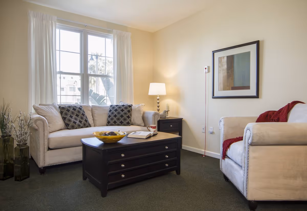 A cozy living room with a beige sofa and armchair, both adorned with decorative pillows and a red throw blanket. A dark wooden coffee table with drawers sits in the center, holding a yellow bowl and an open book. A side table with a lamp and a framed abstract artwork on the wall complete the room. A large window with white curtains lets in natural light.