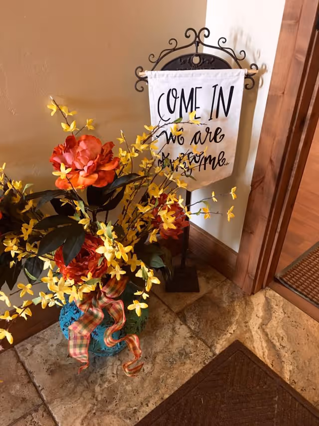 A decorative arrangement with a blue vase holding artificial flowers including large red roses and small yellow blossoms, placed on a tiled floor next to a wooden door. Behind the vase is a small stand with a white fabric sign that reads 'COME IN we are awesome.'