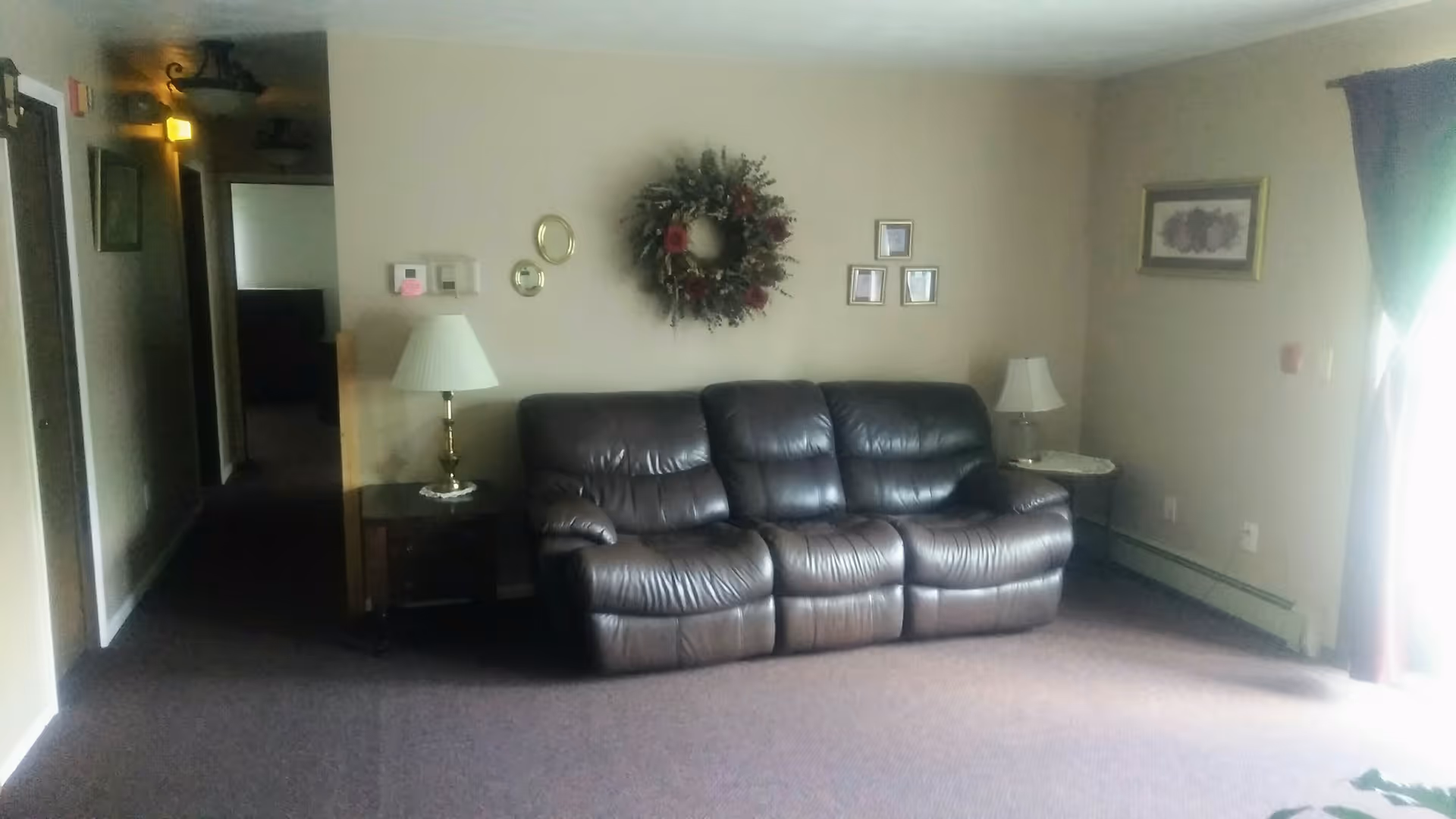 Living room interior with a brown leather three-seat sofa against a wall decorated with a wreath and framed pictures, flanked by side tables and lamps and a hallway to the left.