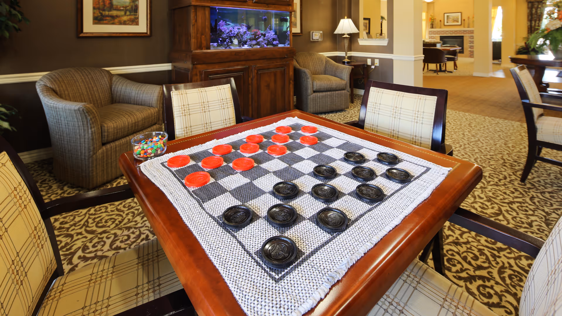 A cozy common area in a senior living facility featuring a wooden table with a checkers game set up on a checkered cloth. Surrounding the table are cushioned chairs with plaid upholstery. In the background, there are comfortable armchairs, a large fish tank on a wooden cabinet, and warm lighting creating a welcoming atmosphere.