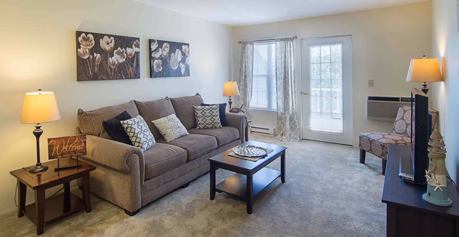 A cozy living room with a beige sofa adorned with decorative pillows, a wooden coffee table, and two side tables with lamps. The room has light-colored walls with two floral paintings above the sofa. There is a window with patterned curtains and a glass door leading outside. A patterned armchair is placed near a TV on a dark wooden stand.