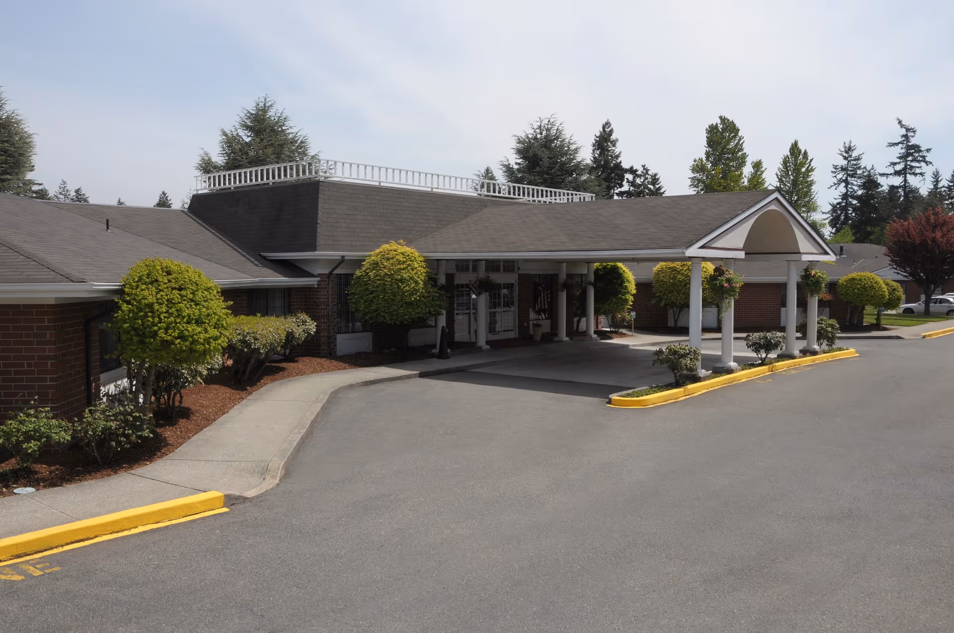 Exterior view of Hallmark Manor showing the entrance with a covered driveway, surrounded by neatly trimmed bushes and trees, under a clear sky.