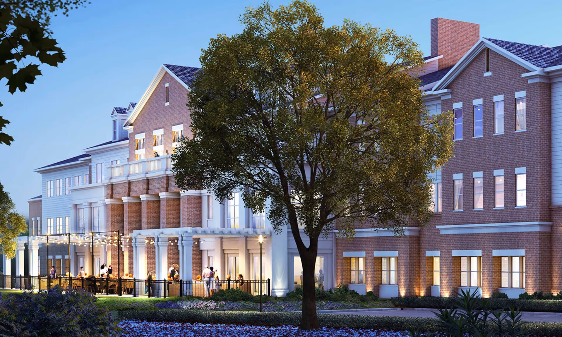 Exterior view of a large brick and white senior living facility building at dusk with outdoor patio seating, string lights, and people gathered outside. A large tree and landscaped garden are in the foreground.
