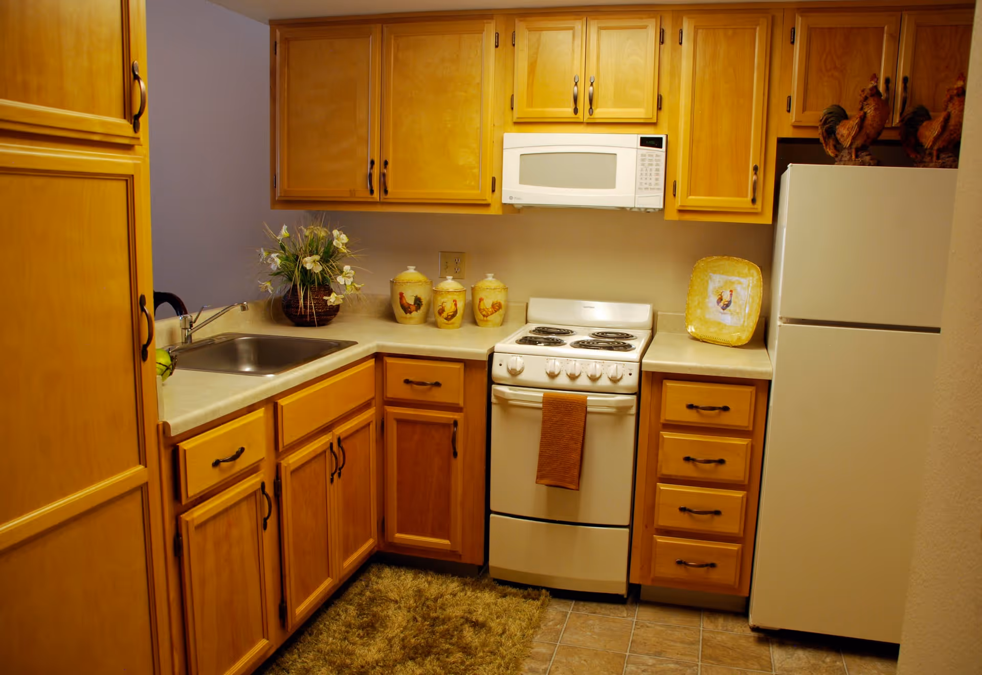 Compact kitchen with honey-colored wooden cabinets, a white stove and microwave, refrigerator, sink, and decorative canisters.