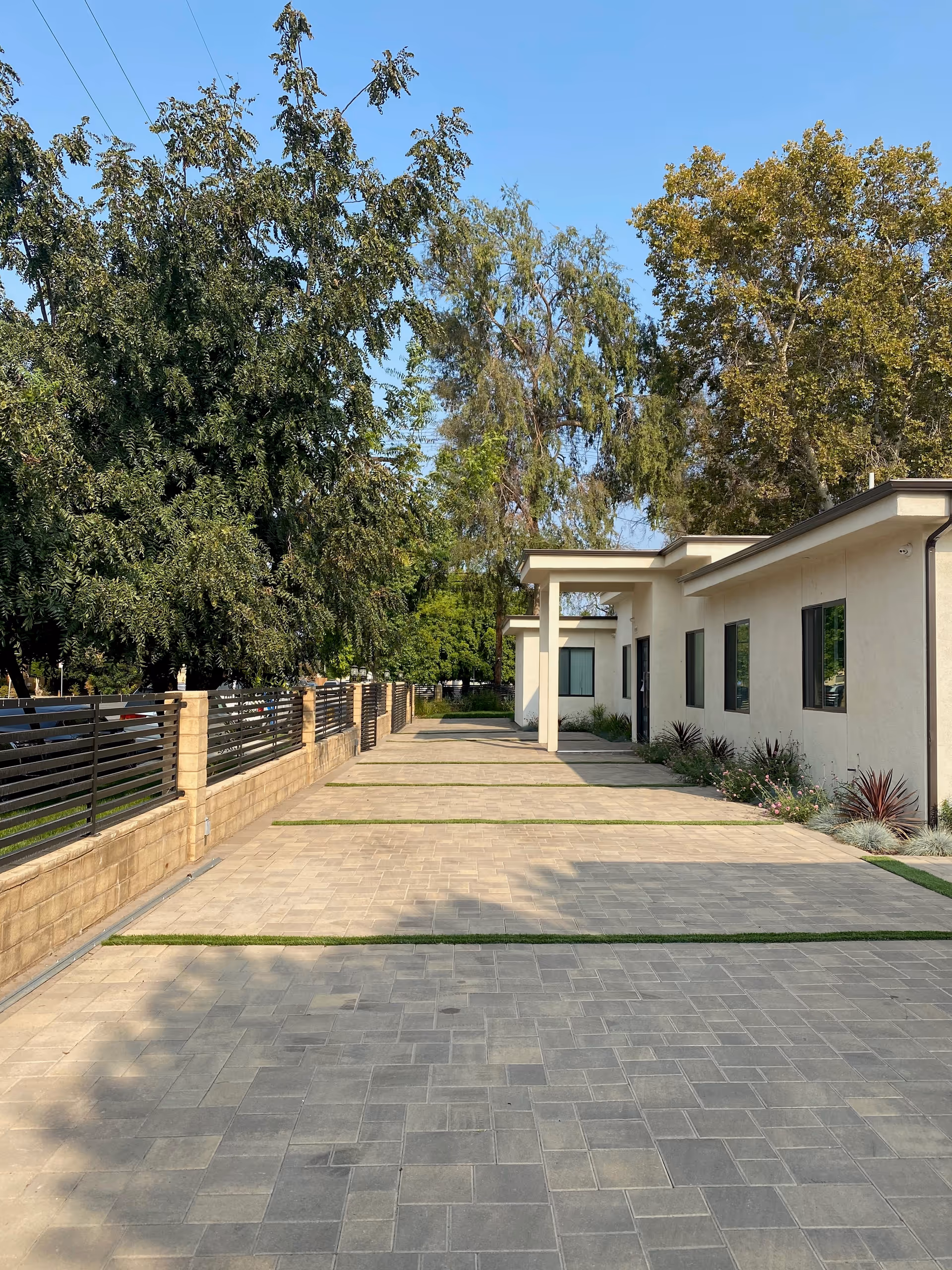 Paved driveway leading to a single-story building with white walls and several windows, bordered by a low brick wall with metal fencing on the left and landscaped plants on the right, under a clear blue sky with tall trees in the background.