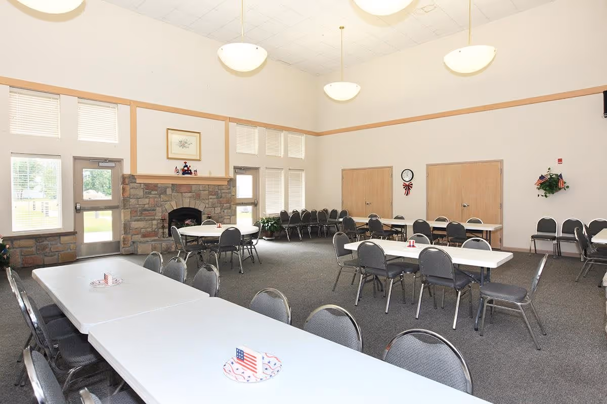 A spacious community dining room with several rectangular and round tables surrounded by gray chairs. The room features a stone fireplace, large windows with blinds, and high ceilings with hanging light fixtures. There are two closed wooden doors on the far wall, a clock, and some patriotic decorations on the tables and walls.