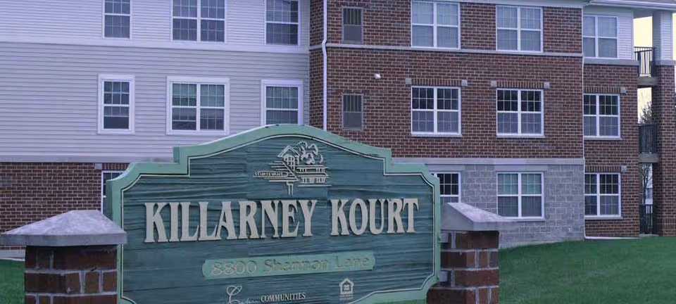 Exterior view of a multi-story residential building with a large green sign in front that reads 'Killarney Kourt' and '8800 Shannon Lane'. The building has a combination of brick and siding with multiple windows visible.