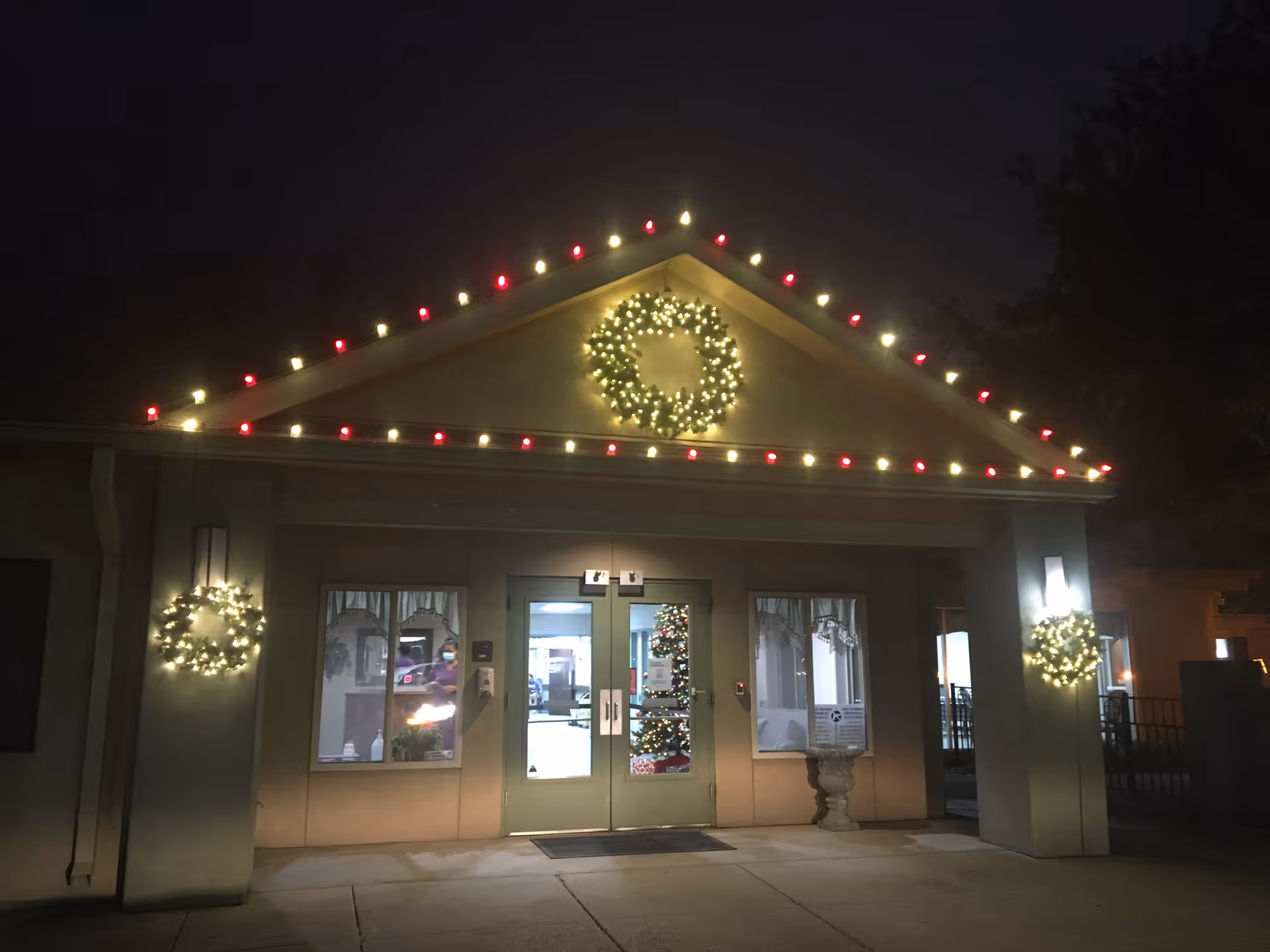 The entrance of Greenbrier Healthcare Center at night, decorated with Christmas lights including a lit wreath above the door and two wreaths on either side of the entrance. The building has double glass doors with a Christmas tree visible inside.