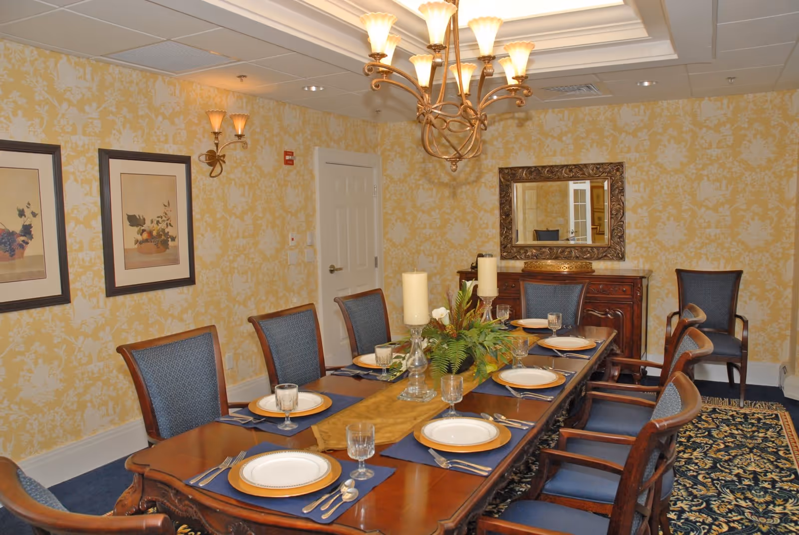 A formal dining room with a long wooden table set for eight people. The table is decorated with a centerpiece of candles and greenery. The room has yellow patterned wallpaper, a chandelier, framed artwork on the wall, and a large ornate mirror above a wooden sideboard.
