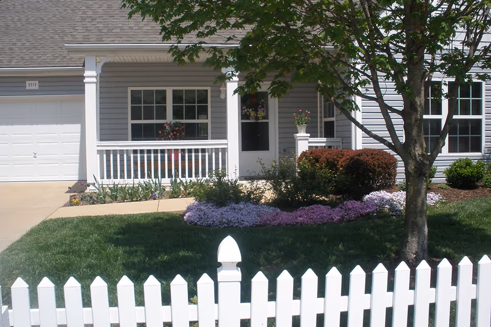 Front view of a single-story house with gray siding, a white picket fence, a small porch with white railings, a white door, and windows. There is a tree and well-maintained garden with green grass, bushes, and purple flowers in the yard.