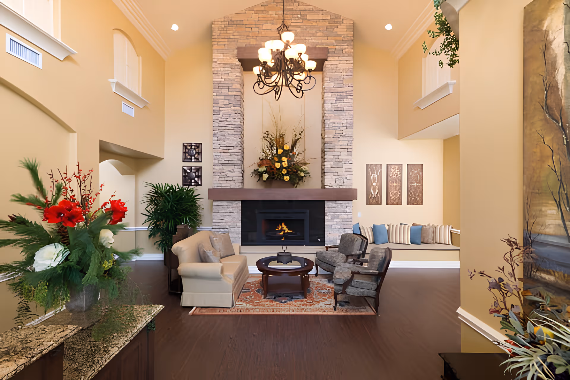 A cozy living room area with a stone fireplace in the center, a chandelier hanging from the ceiling, a beige sofa, two gray armchairs, a round coffee table on a patterned rug, and decorative plants and flowers. The walls are painted a warm beige color, and there are decorative wall hangings and a cushioned bench with pillows along the right wall.