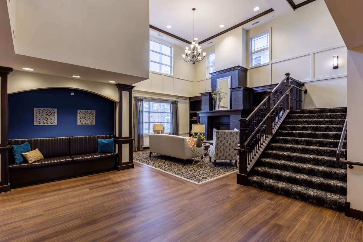 Spacious and elegant living room area with high ceilings, large windows, and a chandelier. The room features a cozy seating arrangement with a white sofa, patterned armchair, and a coffee table on a decorative rug. To the left, there is a built-in bench with cushions against a blue accent wall. A carpeted staircase with dark wooden railings leads to an upper level. The room has wooden flooring and a fireplace with a mirror and decorative vases on the mantel.