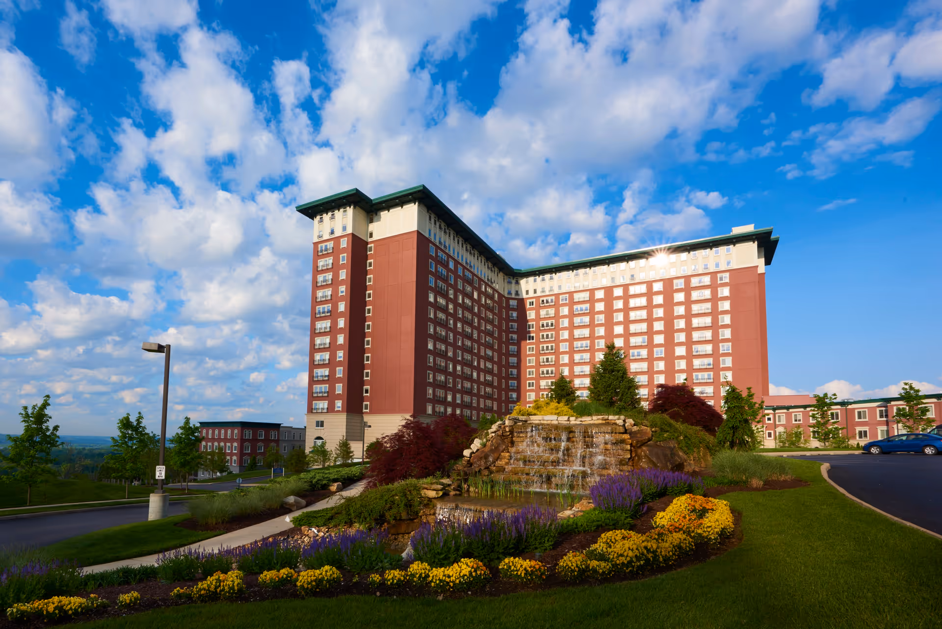 Large multi-story senior living facility building with red and beige exterior under a partly cloudy blue sky. In front of the building is a landscaped area with a rock waterfall feature surrounded by colorful flowers and greenery. A paved driveway curves around the landscaped area.