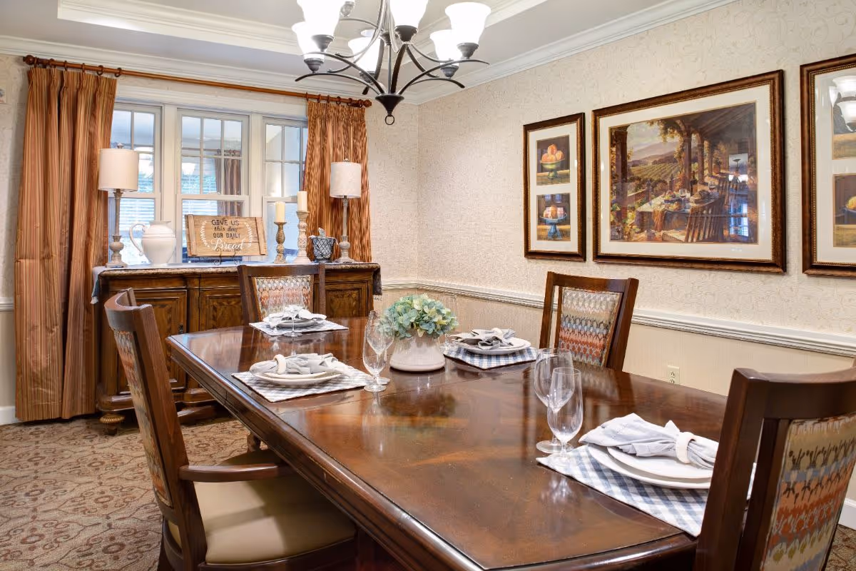 A formal dining room with a polished wooden table set for four, featuring plates, napkins, and glasses. The room has patterned carpet, framed artwork on the walls, a sideboard with decorative lamps and candles, and windows with brown curtains.