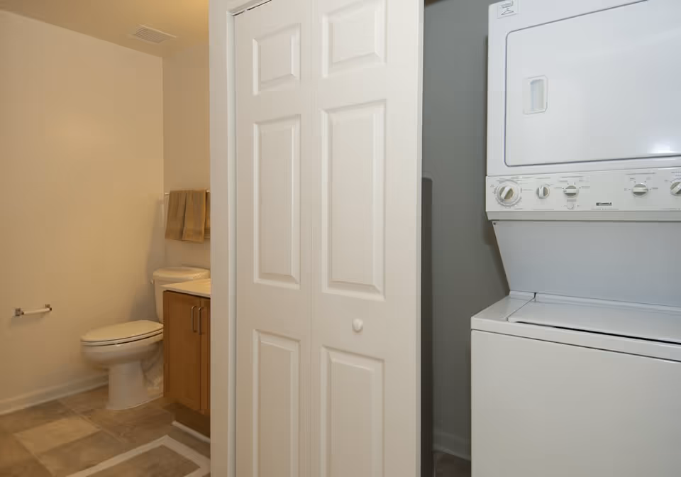 A small bathroom with a toilet, a wooden vanity with a sink, and two beige towels hanging on a towel rack. Adjacent to the bathroom is a laundry area with a stacked white washer and dryer unit partially visible behind white bi-fold doors.
