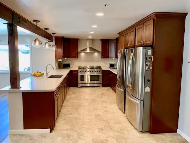 Modern kitchen with dark wood cabinets, stainless steel double-door refrigerator, stainless steel oven with range hood, white countertops, and three pendant lights hanging over the counter. The floor is tiled in a light beige color.