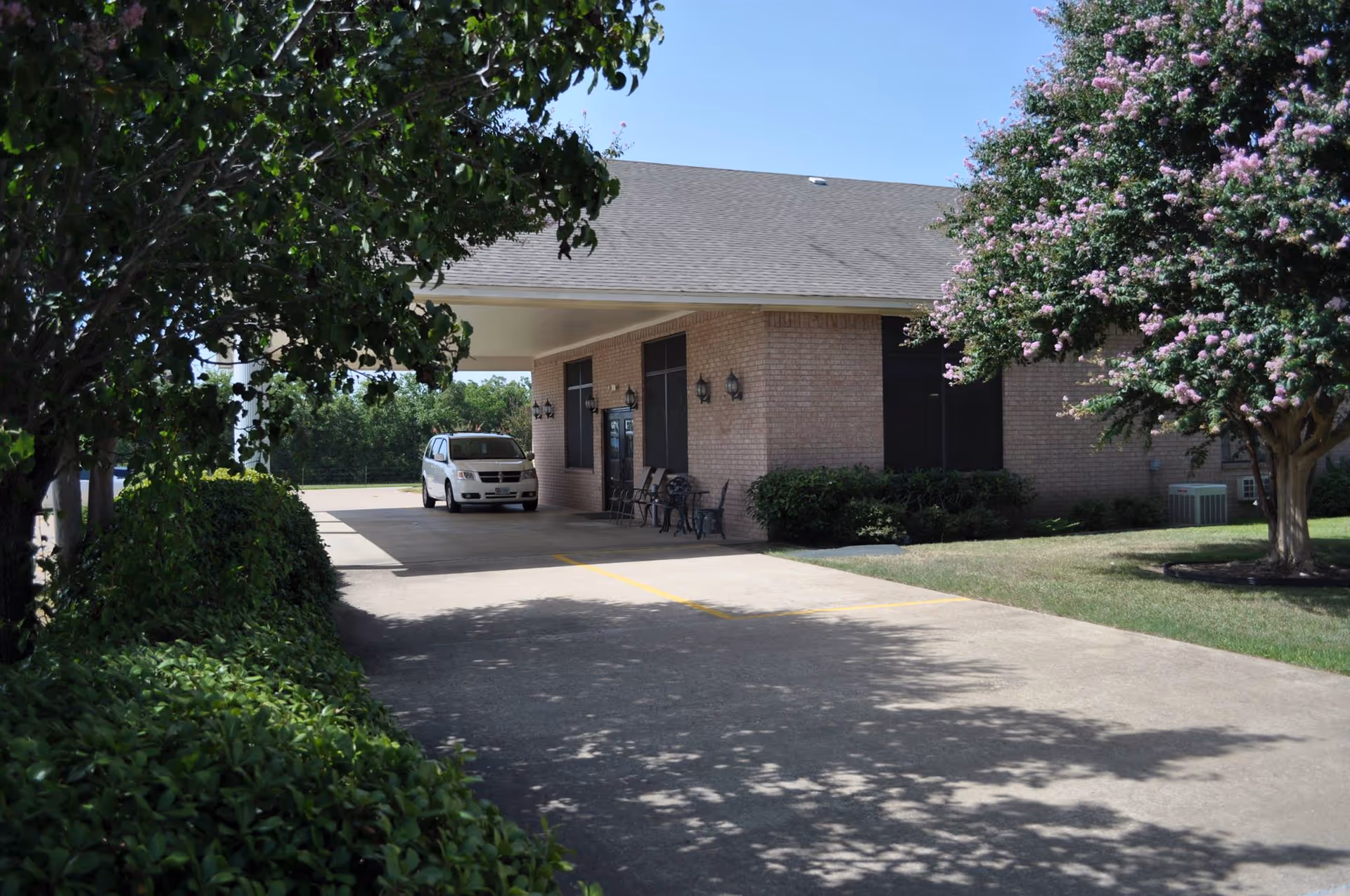 Exterior view of a single-story brick building with a covered driveway. A white vehicle is parked under the covered area. There are green bushes and trees with pink flowers around the building, and the sky is clear and blue.