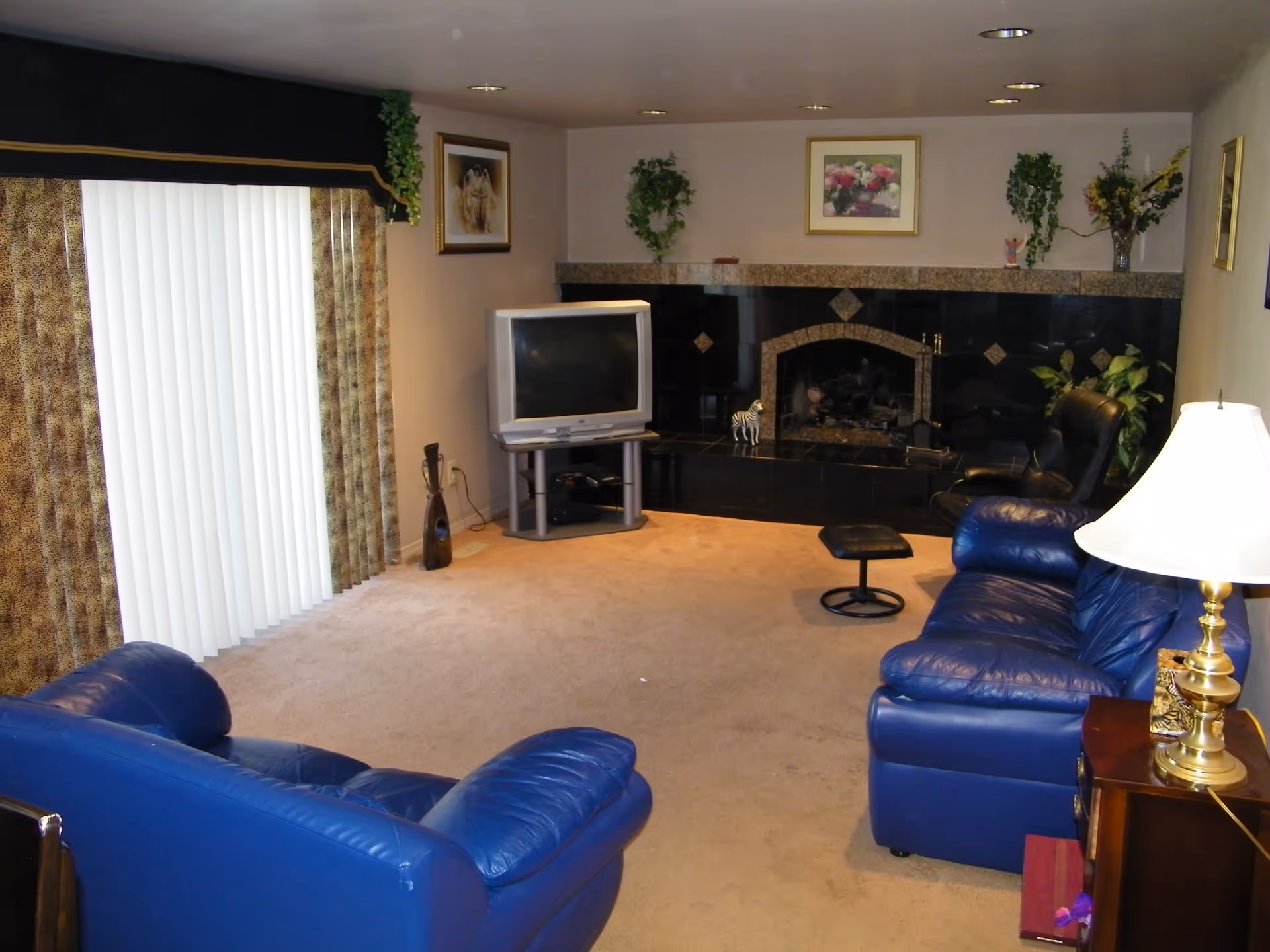A living room with blue leather sofas, a TV on a stand, a black-tiled fireplace, and vertical blinds.