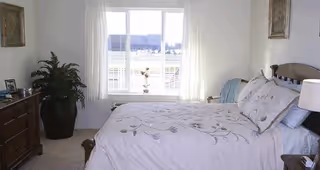 A bright bedroom with a large window letting in natural light. The room features a bed with white floral bedding, a wooden dresser with framed photos, a potted plant, and light-colored curtains. There are framed artworks on the walls.