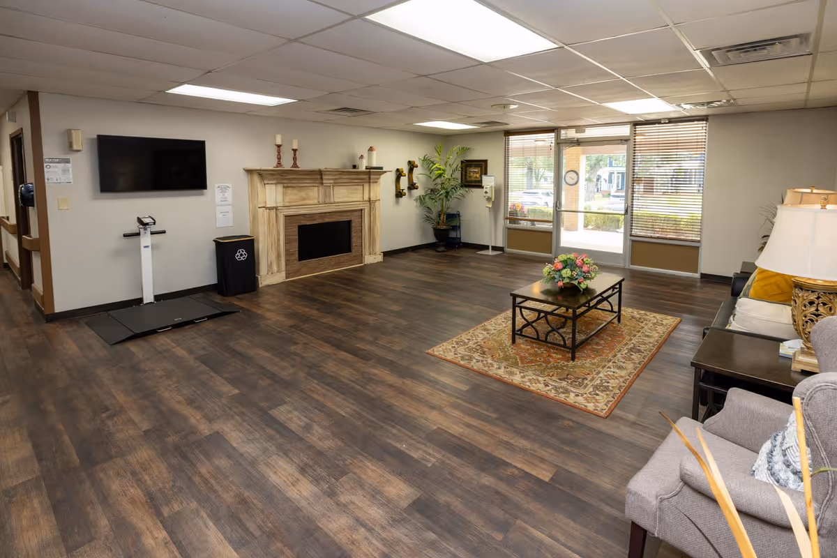 Spacious senior living common lounge with seating, a coffee table on an area rug, decorative fireplace, wall-mounted TV, and entrance doors.