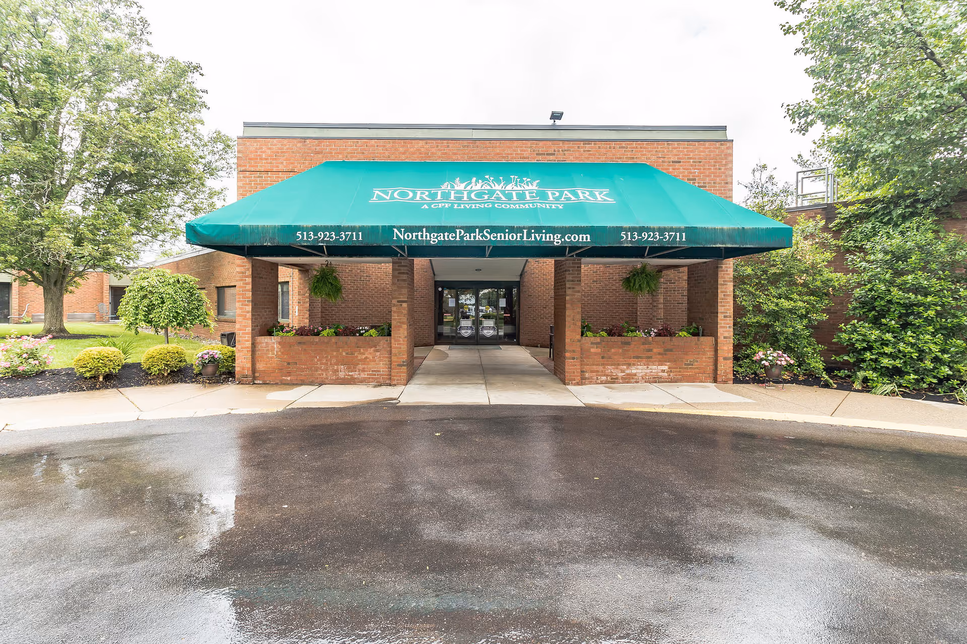 Front entrance of a brick senior living building with a green awning and a wet circular driveway.