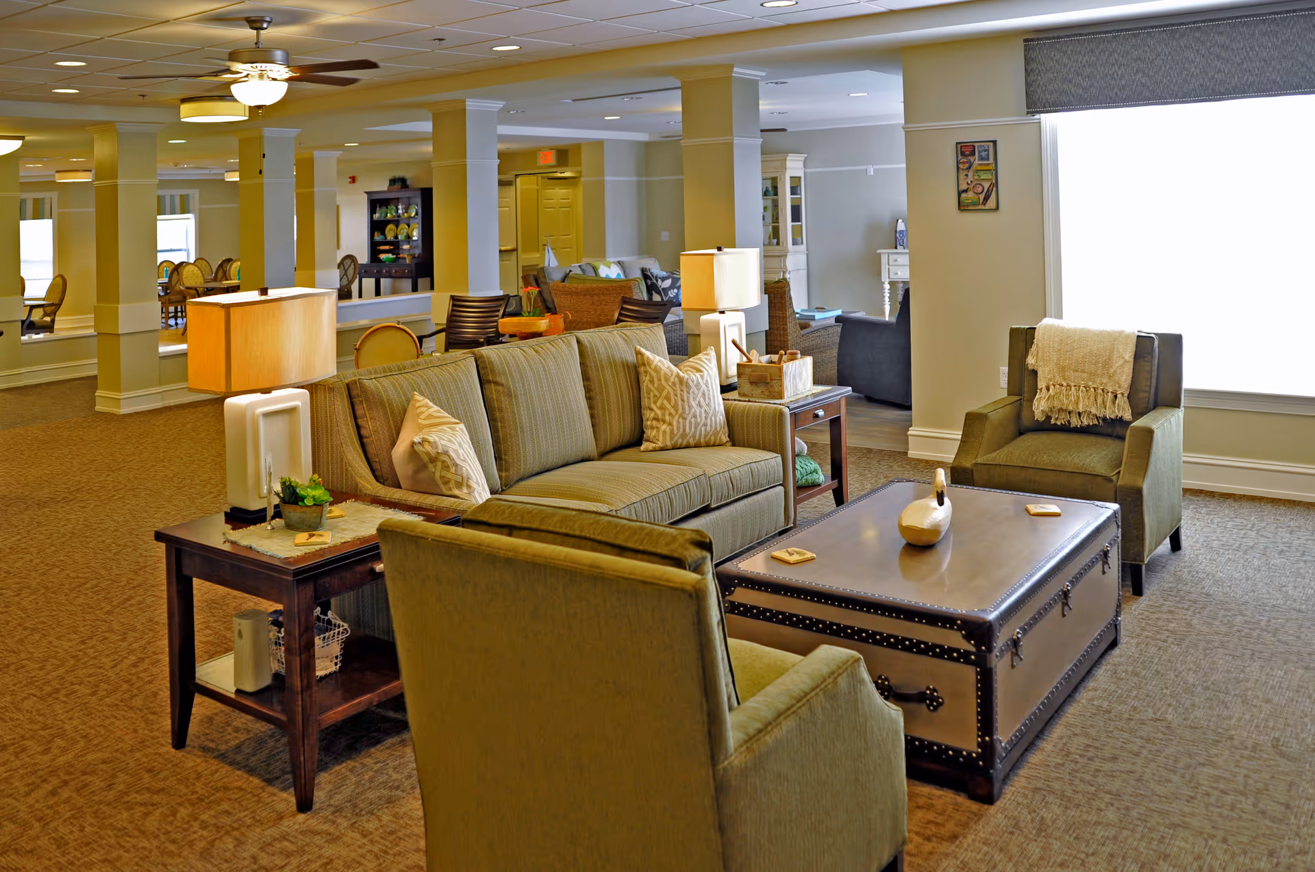 A cozy senior living facility common area with a beige sofa, two green armchairs, a large trunk-style coffee table, and side tables with lamps. The room has carpeted floors, ceiling fans, and large windows letting in natural light. In the background, there are more seating areas and decorative elements.