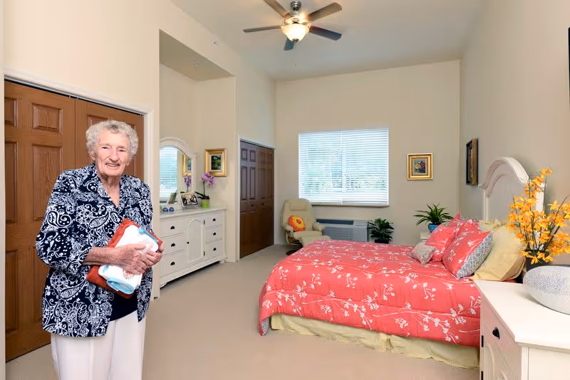 A senior woman standing in a spacious bedroom with beige walls and carpet. The room features a bed with a red floral comforter, a white dresser with a mirror, a beige armchair near a window with blinds, and decorative plants and flowers.