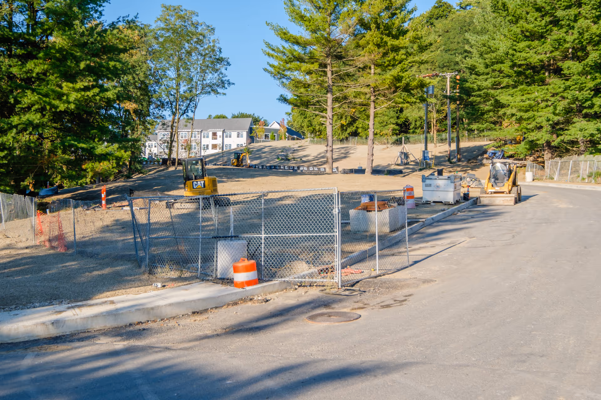 Fenced construction site with small excavators and traffic barrels beside a curved road, with trees and a residential building in the background.