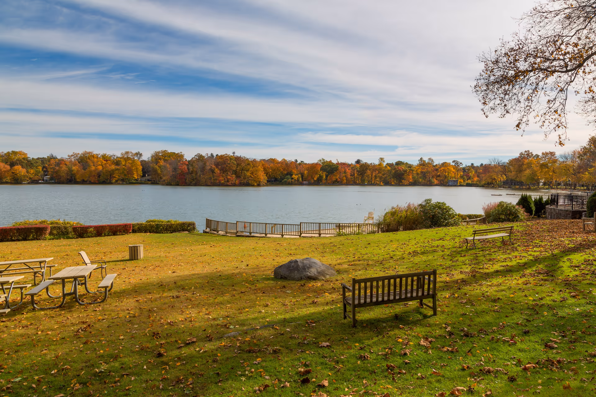 A peaceful outdoor area with green grass, scattered fallen leaves, wooden benches, and picnic tables overlooking a calm lake. Trees with autumn-colored foliage surround the lake under a partly cloudy sky.